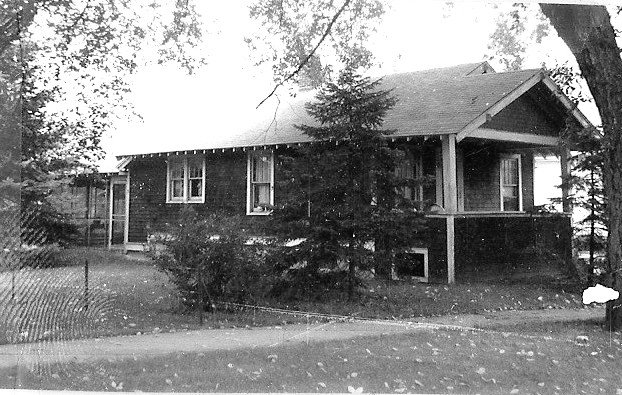 Black-and-white photo of a single-story bungalow with a covered front porch, evergreen trees in front, and a grassy yard.