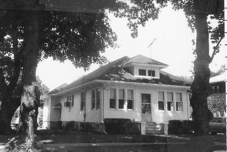 Black-and-white photo of a one-story bungalow with an enclosed front porch, small front steps, and two large trees framing the house.