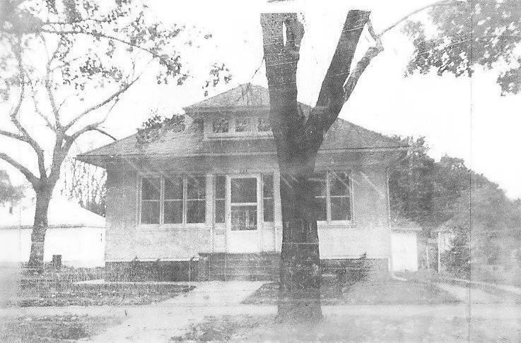 Black-and-white photo of a single-story bungalow with a central front door and a large tree trunk in the foreground.