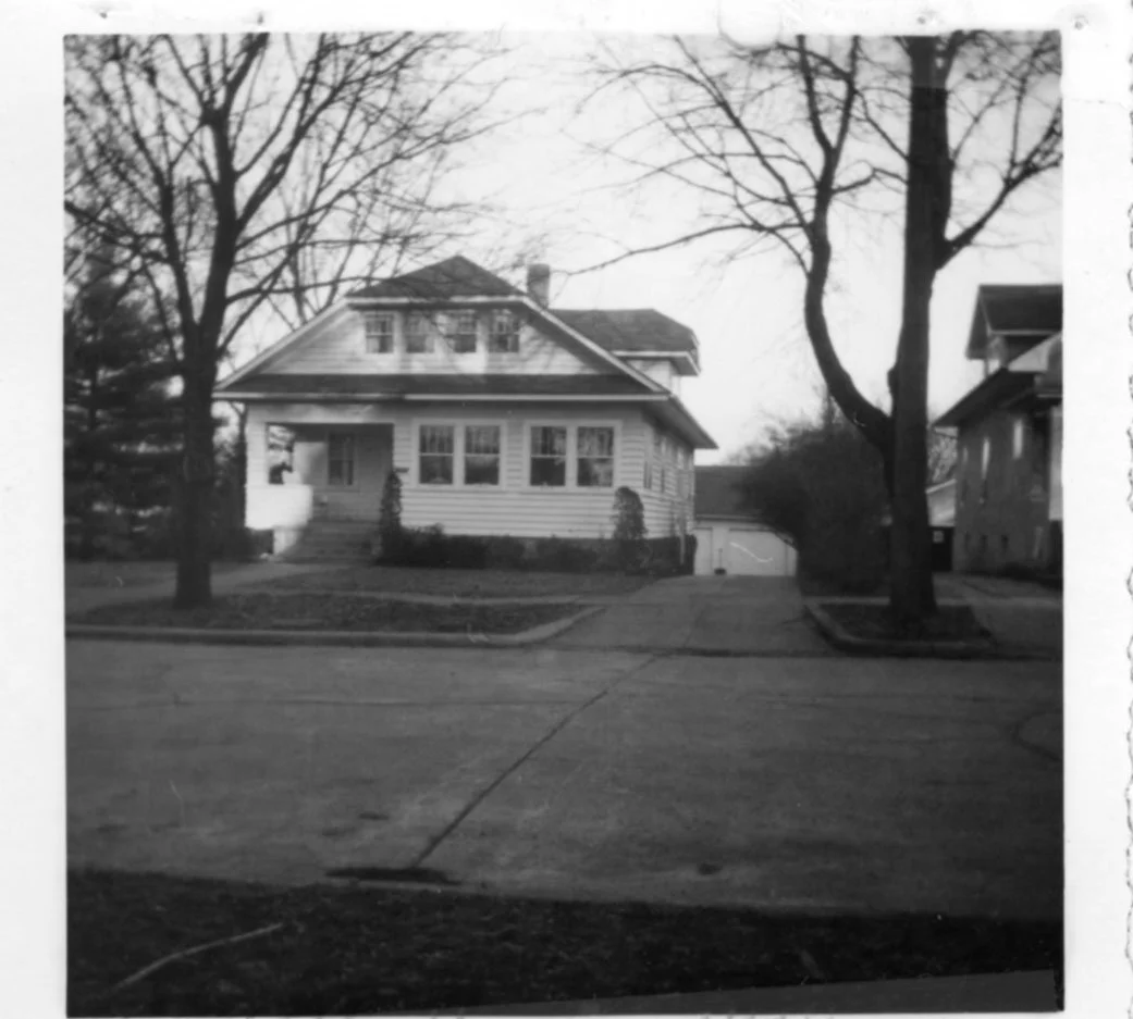 Black-and-white photo of a two-story bungalow with a front porch, leafless trees framing the yard, a driveway leading to a detached garage, and neighboring houses.