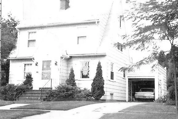 Black-and-white photo of a two-story bungalow with a front porch, leafless trees framing the yard, a driveway leading to a detached garage, and neighboring houses.