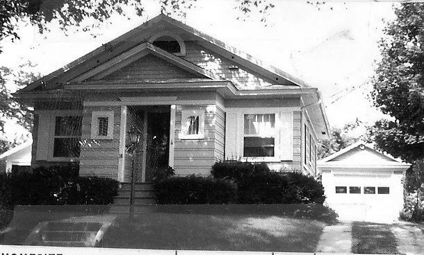 Black-and-white photo of a single-story bungalow with a small front porch, shrubs lining the facade, a detached garage to the right, and a lawn and trees in front.