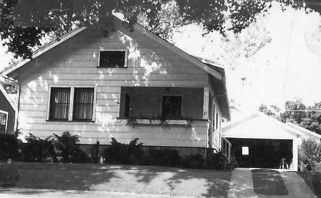 Black-and-white photo of a single-story bungalow with a small covered front porch and a detached garage to the right, tree shadows falling across the house.