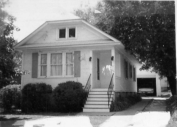 Black-and-white photo of a single-story bungalow with front steps and iron railings, shrubs along the porch, and a driveway leading to a detached garage with a car inside.