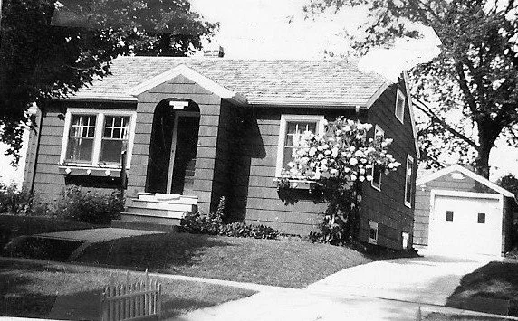 Black-and-white photo of a small single-story house with a front porch, flower boxes under the windows, a sloped lawn, and a detached garage to the right.