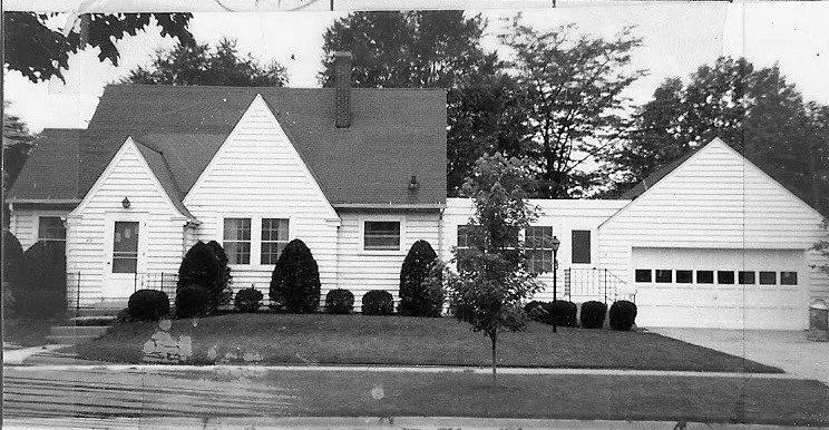 Black-and-white photo of a single-story suburban house with steep gables, front shrubs and small tree, and an attached two-car garage.