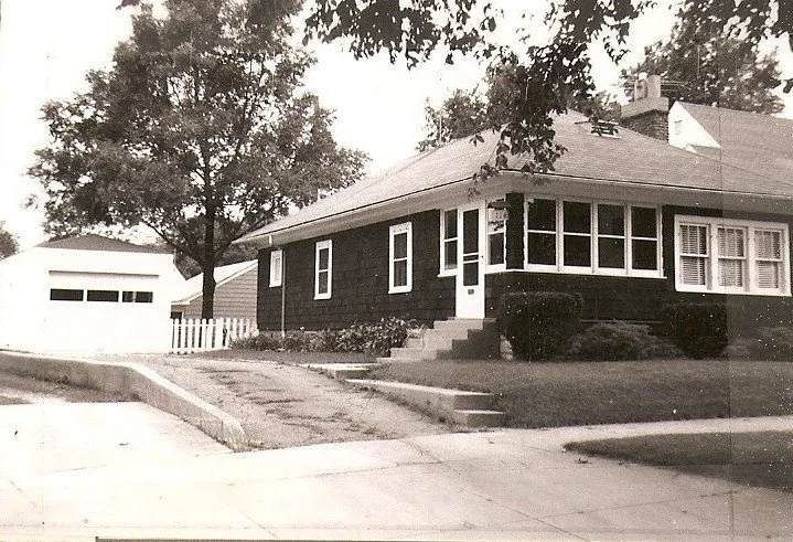 Black-and-white photo of a single-story bungalow with an enclosed front porch, concrete steps, shrubs and a driveway leading to a detached garage, framed by trees.