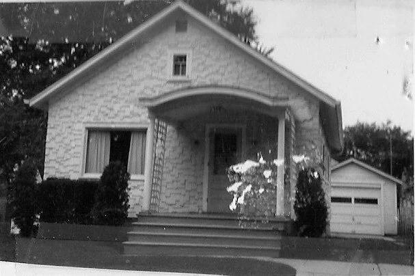 Black-and-white photo of a small bungalow with a covered front porch and steps, shrubs flanking the entrance, and a detached garage to the right.