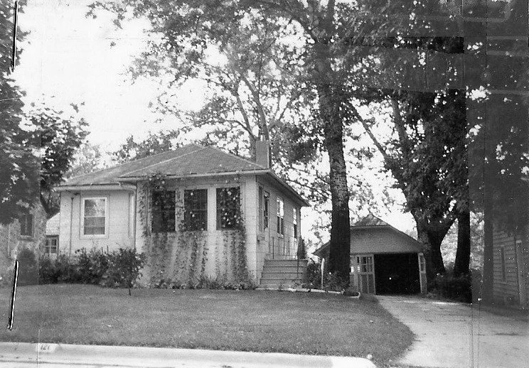 Black-and-white photo of a small one-story house with a front porch, lawn, driveway, detached garage, and large trees.