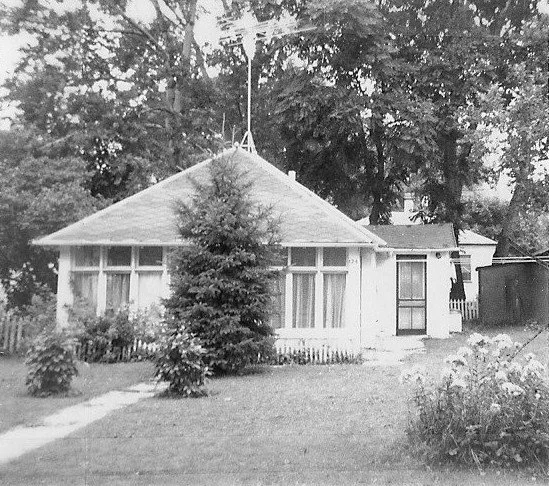 Black-and-white photo of a small single-story bungalow with large front windows, an evergreen tree and walkway in the yard, and tall trees behind.