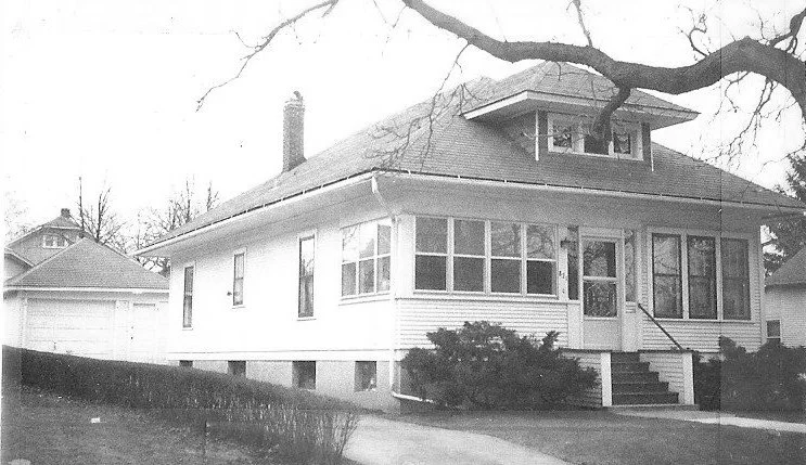Black-and-white photo of a single-story bungalow with an enclosed front porch, front steps, detached garage to the left, and bare tree branches overhead.