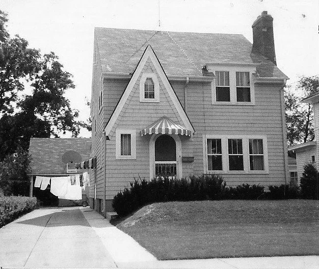 Black-and-white photo of a two-story house with a steep front gable and striped awnings, driveway and carport with laundry hanging on a clothesline, and a neatly trimmed front lawn.