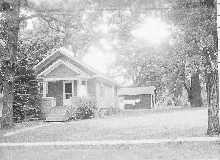 Black-and-white photo of a small bungalow with a front porch and detached garage surrounded by large trees on a grassy lot.