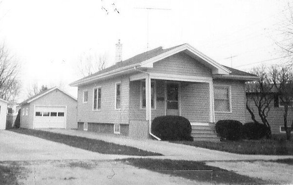 Black-and-white photo of a single-story bungalow with a small covered front porch, shrubs, driveway, and a detached two-car garage in the background.