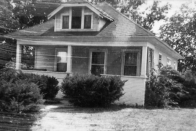 Black-and-white photo of a single-story bungalow with a covered front porch, dormer window, and shrubs in the front yard.