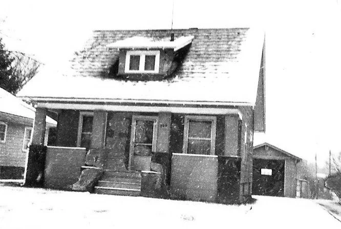 Black-and-white photo of a small two-story house with a covered front porch, snow on the roof and ground, and a detached garage to the right.