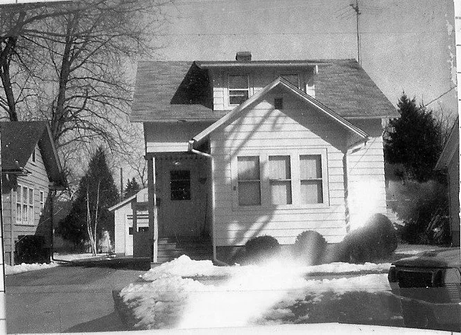 Black-and-white photo of a small two-story house with a snowy front yard, bare trees, and a car parked at the right edge.
