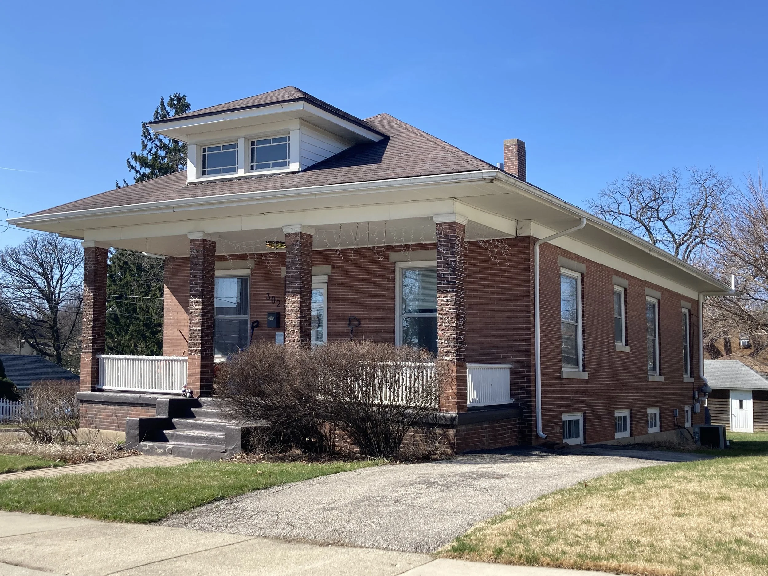 Brick two-story house with front porch, dormer window, chimney, and clear blue sky.