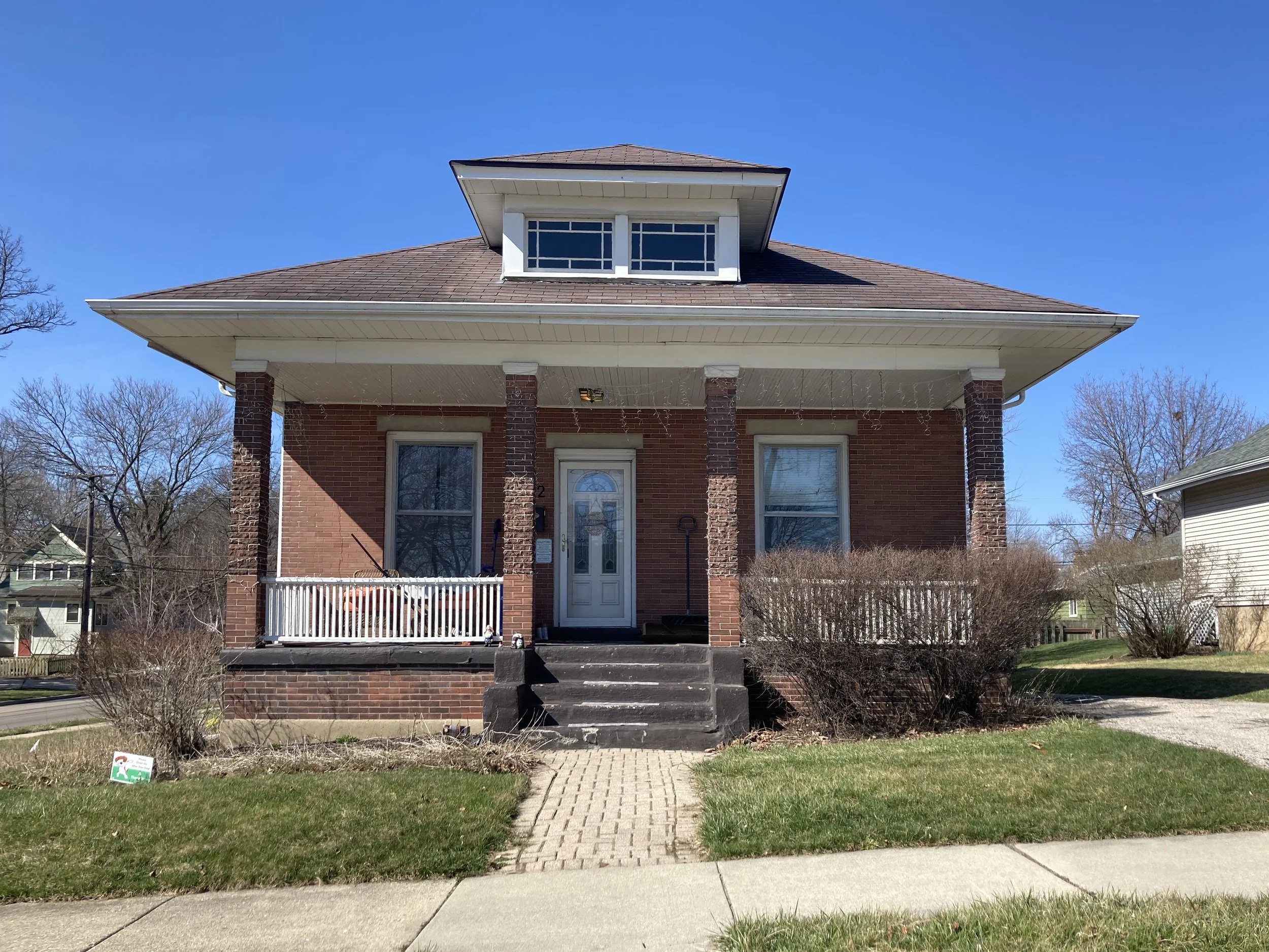 Front view of a brick house with porch, steps, dormer window, and clear blue sky.