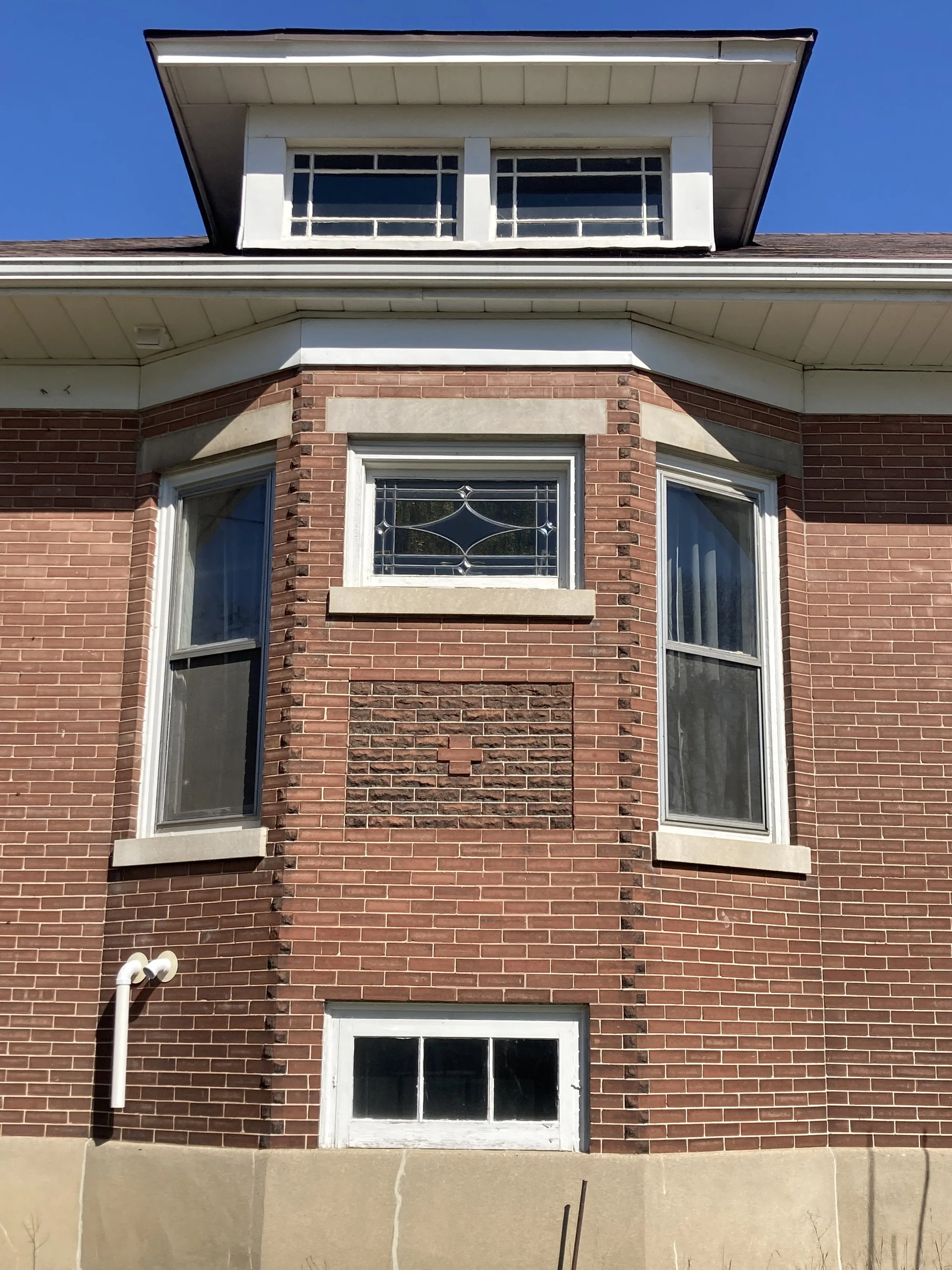  Brick house front with bay windows, a small stained-glass window, and a dormer under a blue sky.
