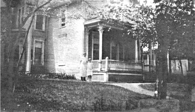 Black-and-white photo of an old house with a covered porch and two people standing on the lawn.