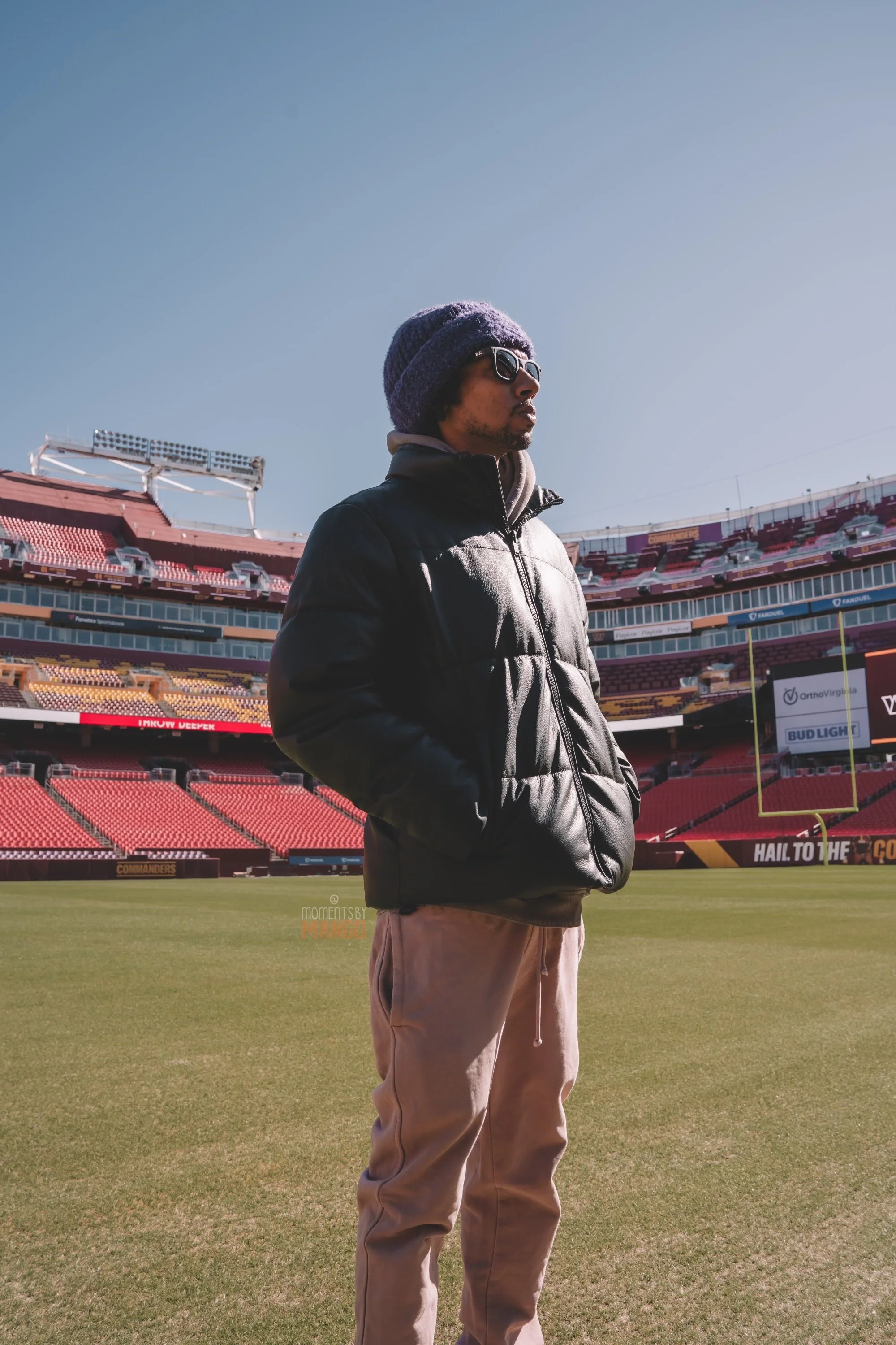 A man wearing sunglasses, a knit hat, a jacket, and khaki pants stands on the field of a football stadium with red seats and blue sky in the background.