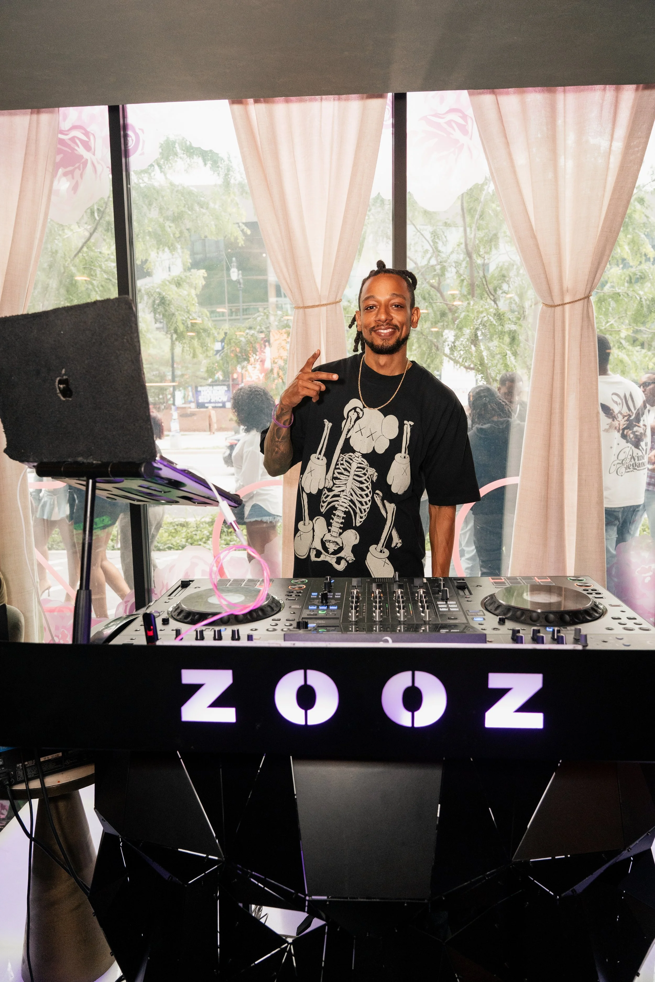 A man with dreadlocks and a goatee, wearing a black T-shirt with a skeleton design, standing behind DJ equipment in a room with large windows, beige curtains, and outdoor trees visible through the windows.