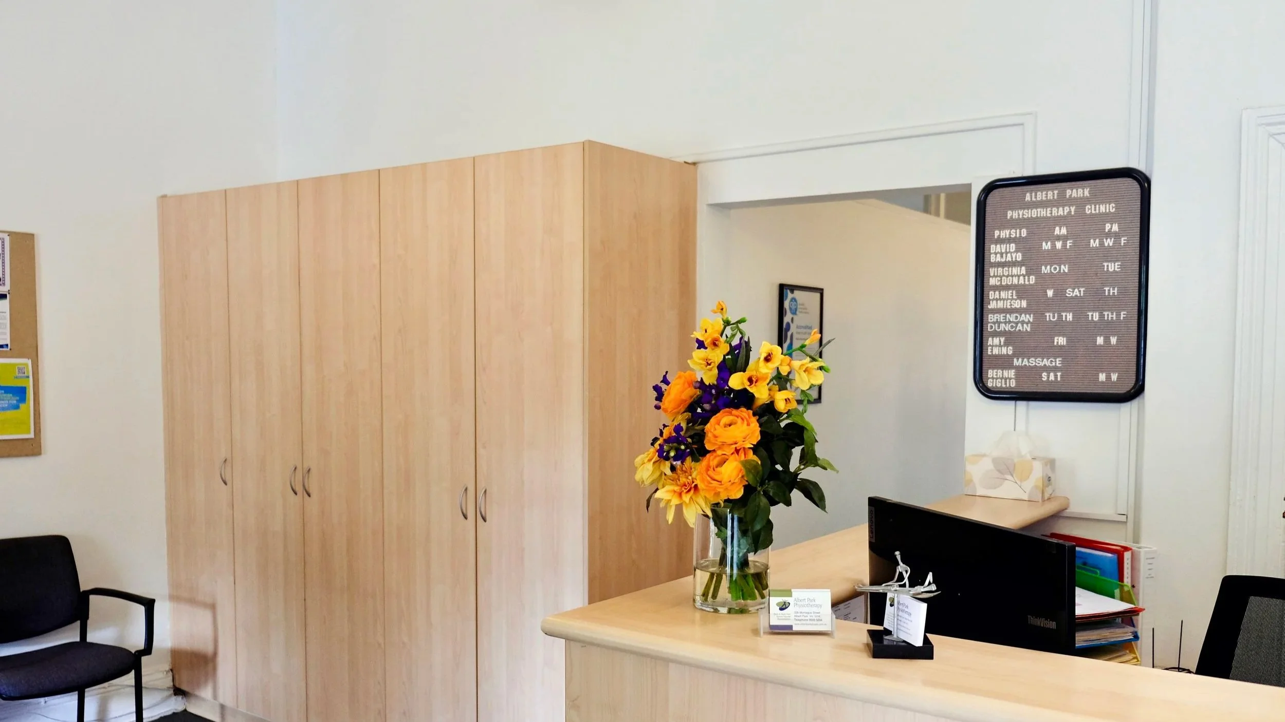 Reception area of a physiotherapy clinic with black schedule board on the wall listing staff and hours.