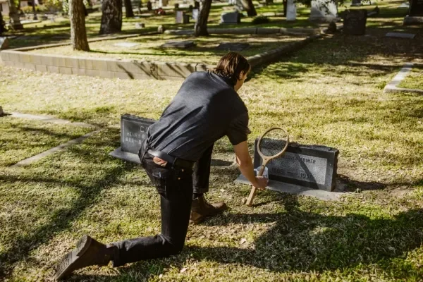 Man rests old tennis racket against a tombstone in cemetery