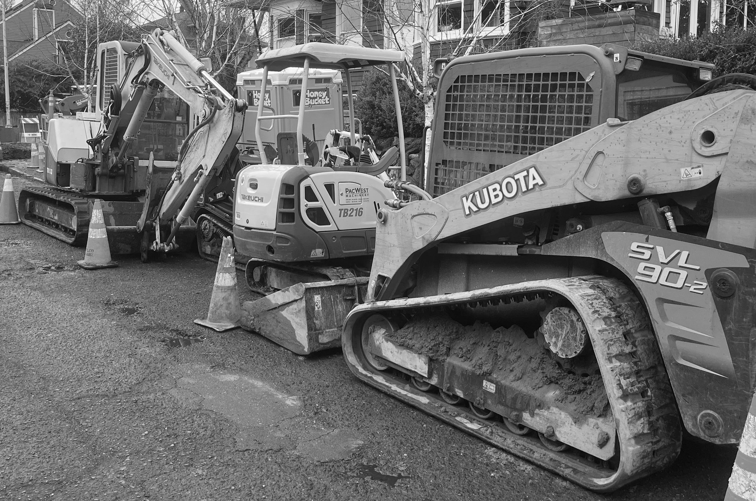 Construction equipment including a Kubota SVL 90-2 compact track loader, a Takeuchi TB216 mini excavator, and other machinery, lined up on a street with construction cones, in a residential neighborhood.