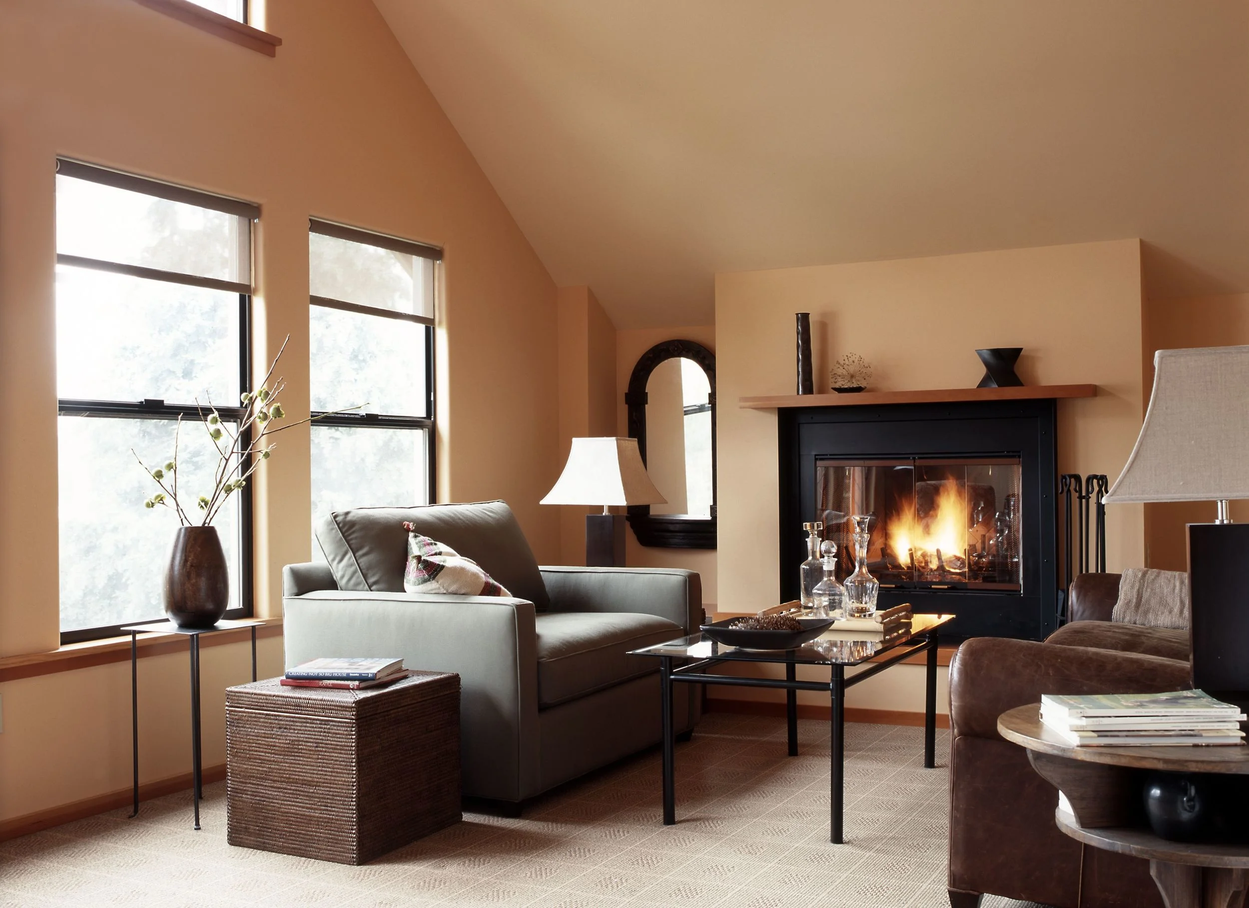 Living room with beige walls, large windows, a sofa with a pillow, a side table with a vase and branches, a coffee table with glass bottles, and a fireplace with a fire burning.