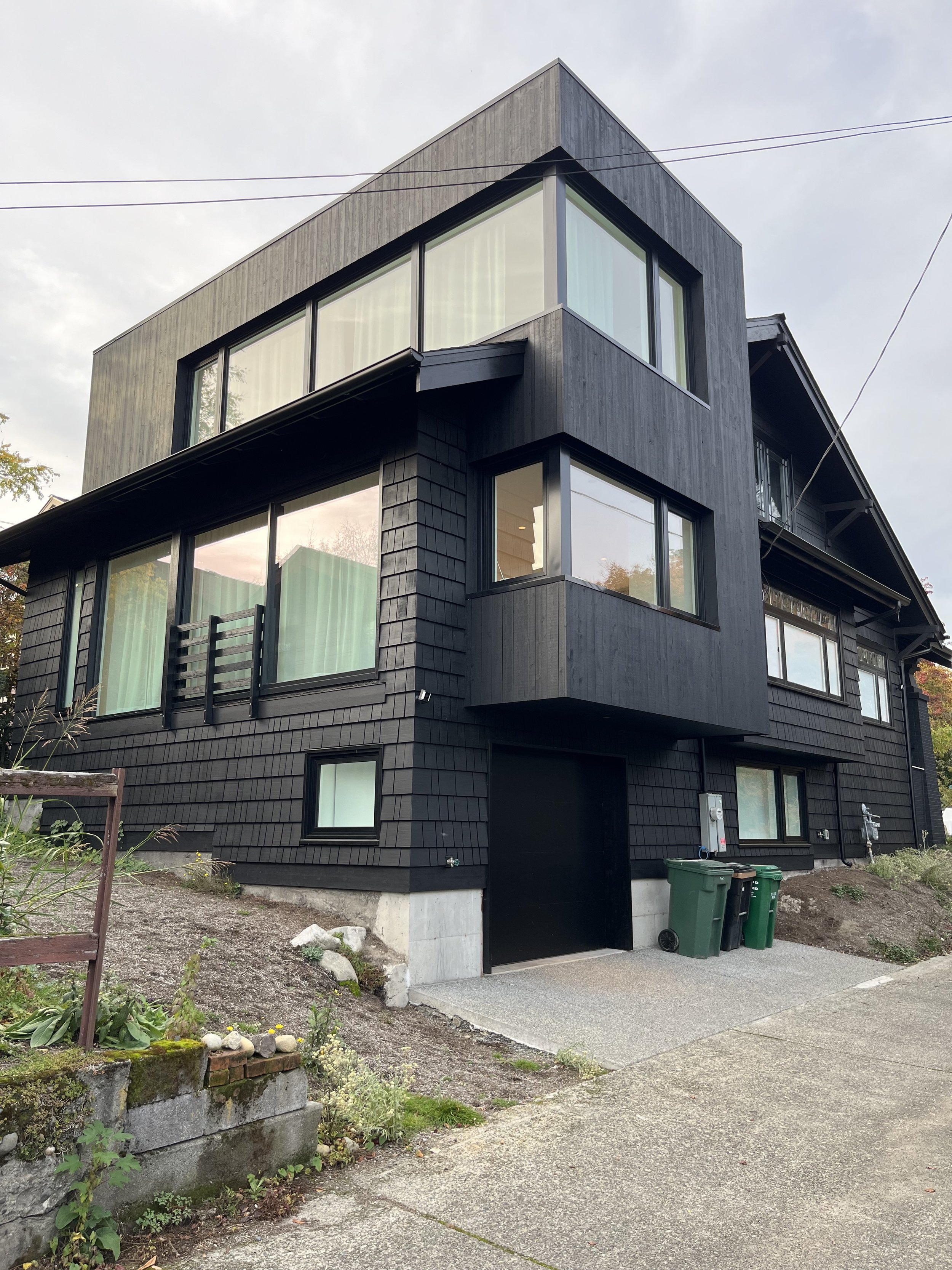 Modern multi-story black house with large windows, gray accents, and a small garden area in the front.