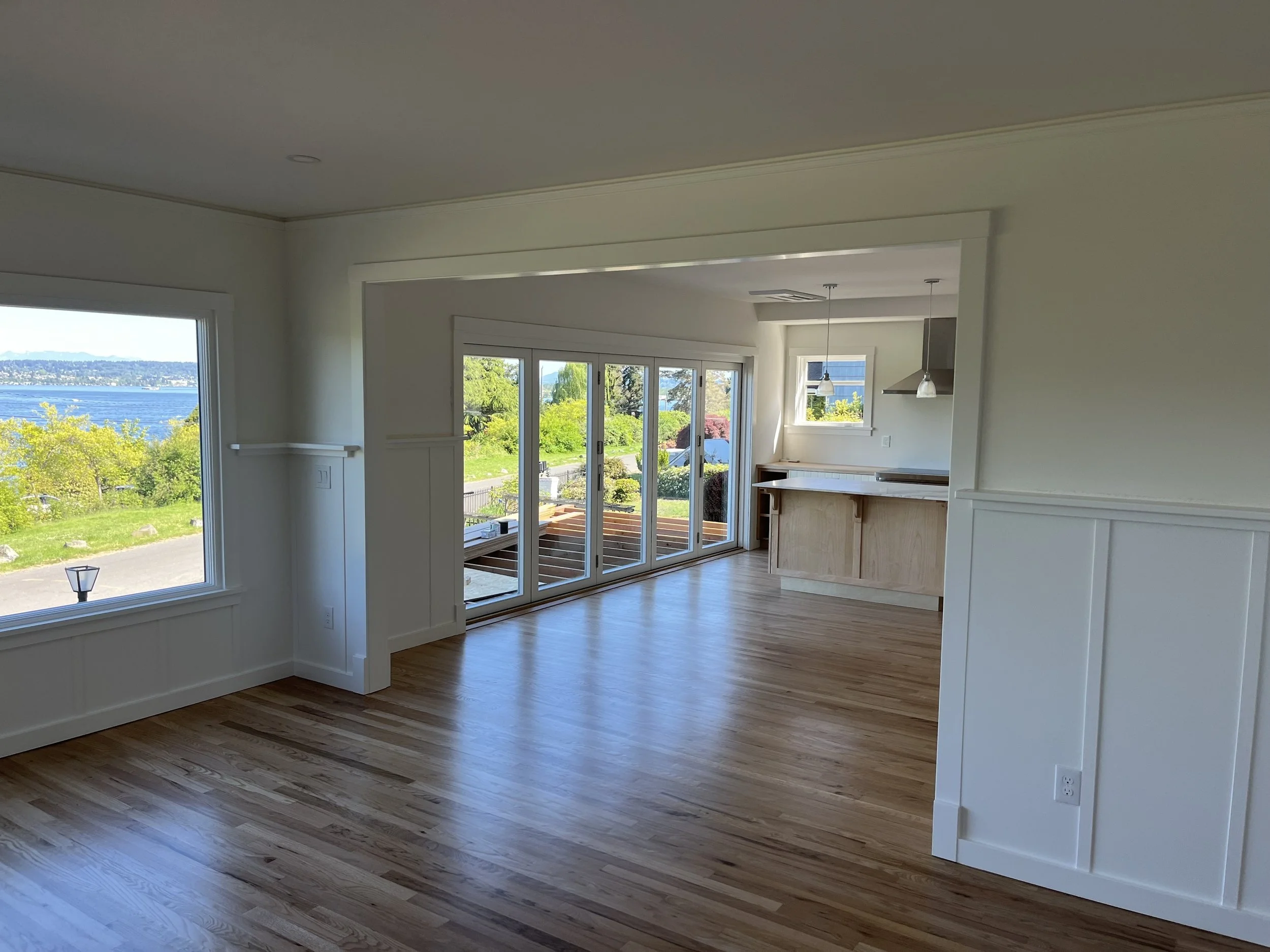 Empty living room with large windows showing a river view, opens to a kitchen area with hanging lights and a small window, hardwood floors, and white walls.