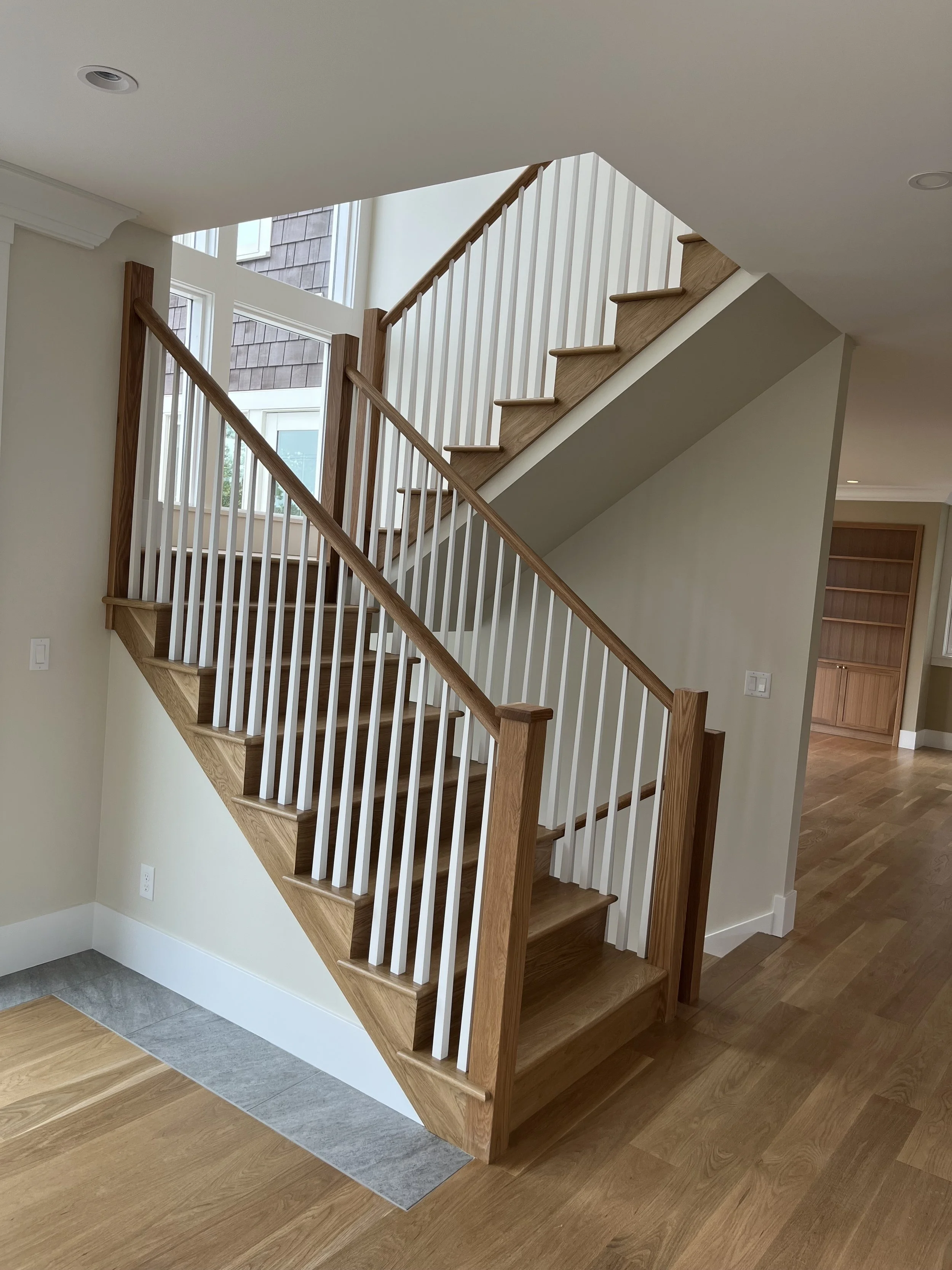 Wood staircase with white spindles and a wooden handrail leading to an upper floor in a modern home with large windows and hardwood floors.