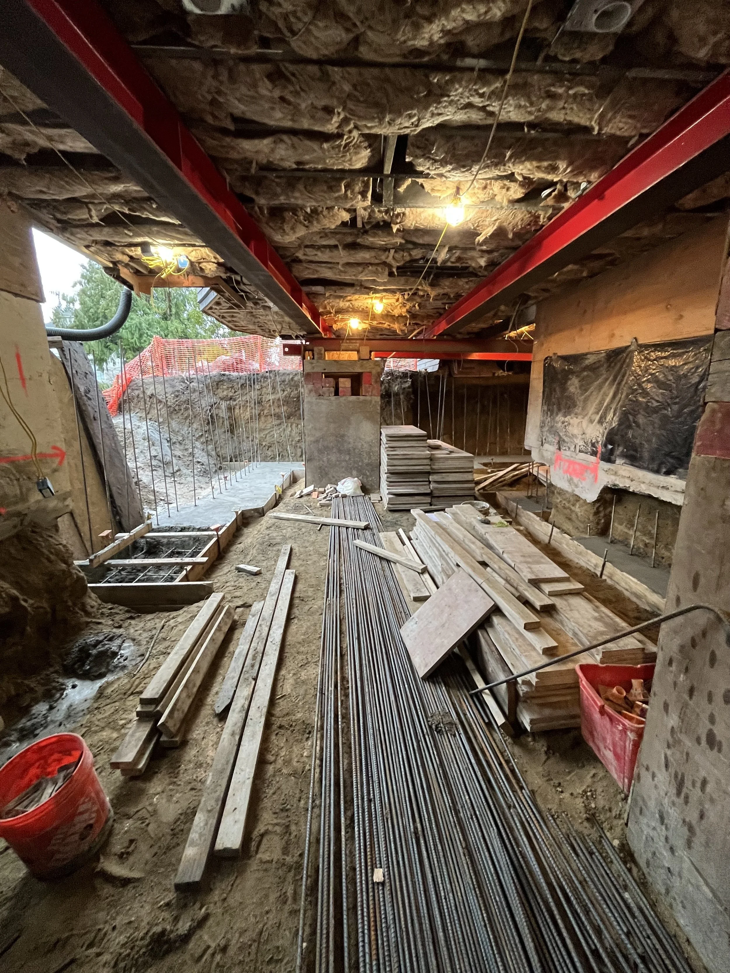 Construction site with building foundation, wooden and metal reinforcement bars, construction materials, and partial ceiling insulation, with orange safety fencing outside in the background.