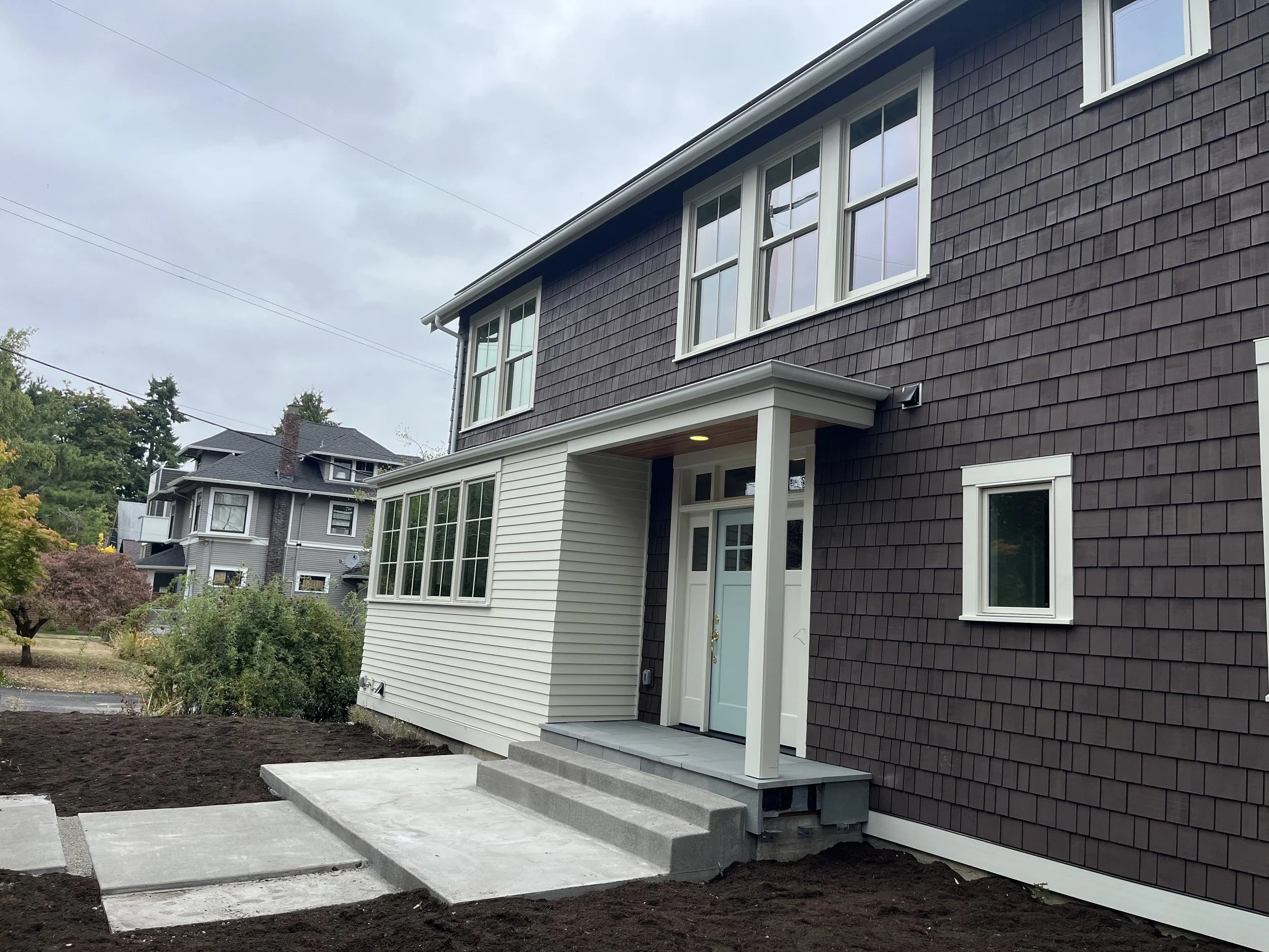Newly constructed two-story house with a front porch, gray shingles on upper level, white siding on lower level, and concrete steps leading to the front door, in a neighborhood with other houses and trees.