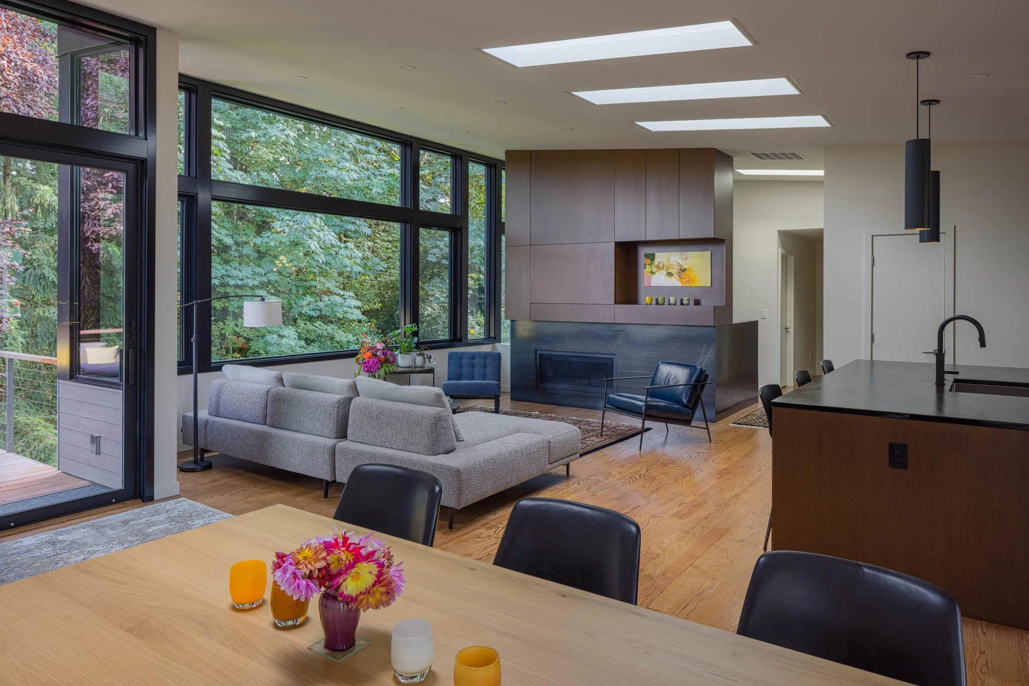 Open-concept living room and dining area with large windows showing greenery outside. Modern furniture including a gray sofa, black and blue chairs, and a wooden dining table with colorful flowers and candles. Kitchen island with black countertop and elegant pendant lighting.