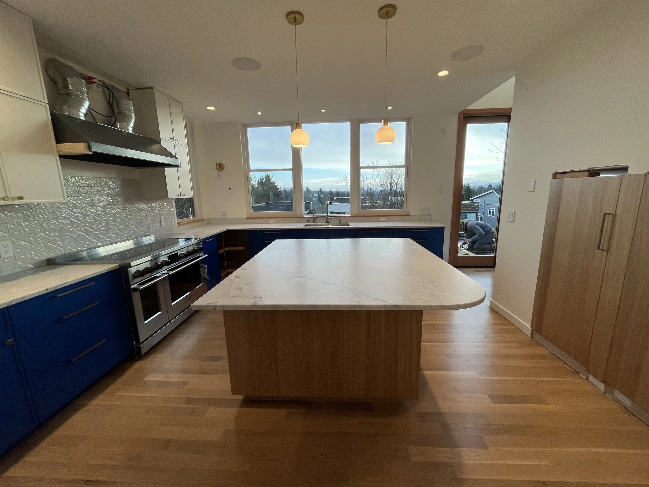 Modern kitchen with white and blue cabinets, a large marble island, stainless steel stove, and large windows overlooking the outdoors.