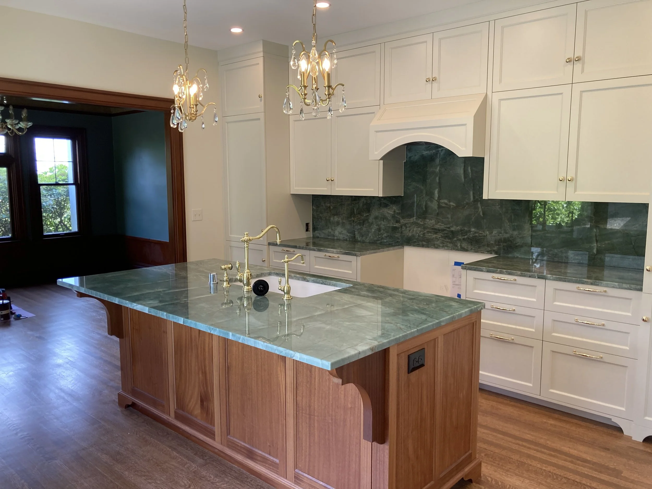 Kitchen with white cabinets, green marble backsplash, green marble countertop on the island, brass fixtures, and wooden flooring. Two chandeliers hang from the ceiling.