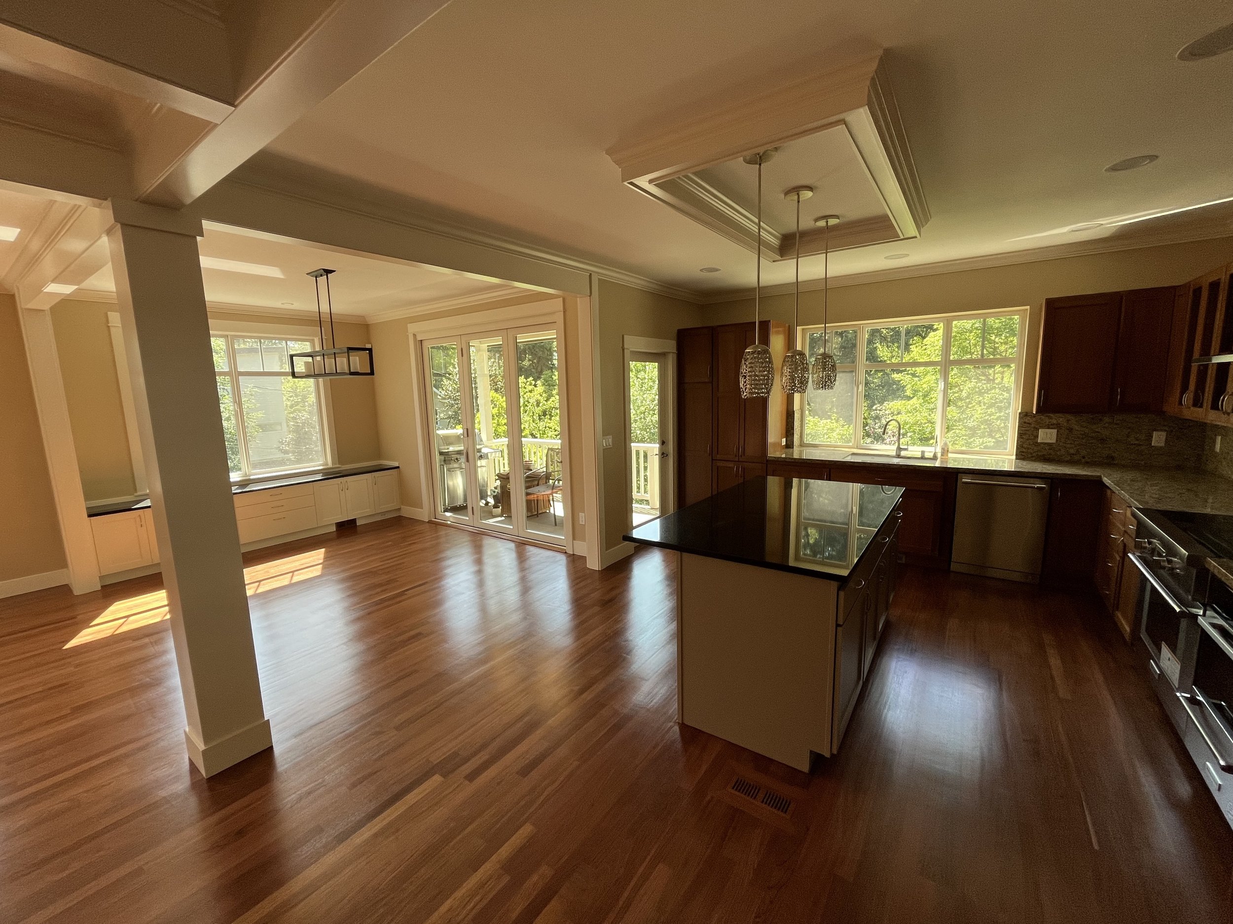 Empty kitchen and dining area with hardwood floors, large windows, and a sliding glass door leading to a balcony. The kitchen features granite countertops, dark wood cabinets, and an island with pendant lighting.