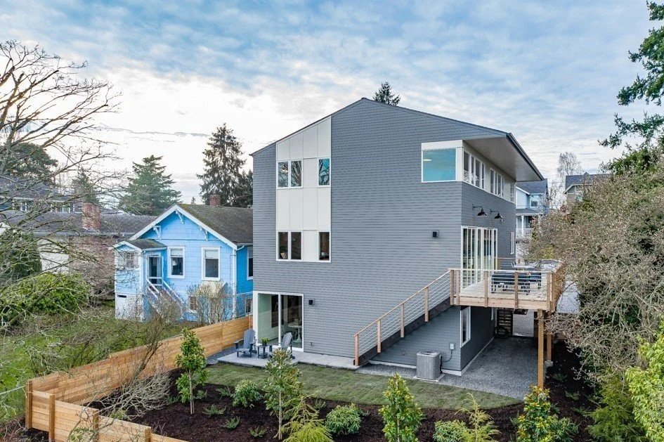 Modern gray house with large windows and a wooden deck, neighboring a small blue house, surrounded by trees and a wooden fence.