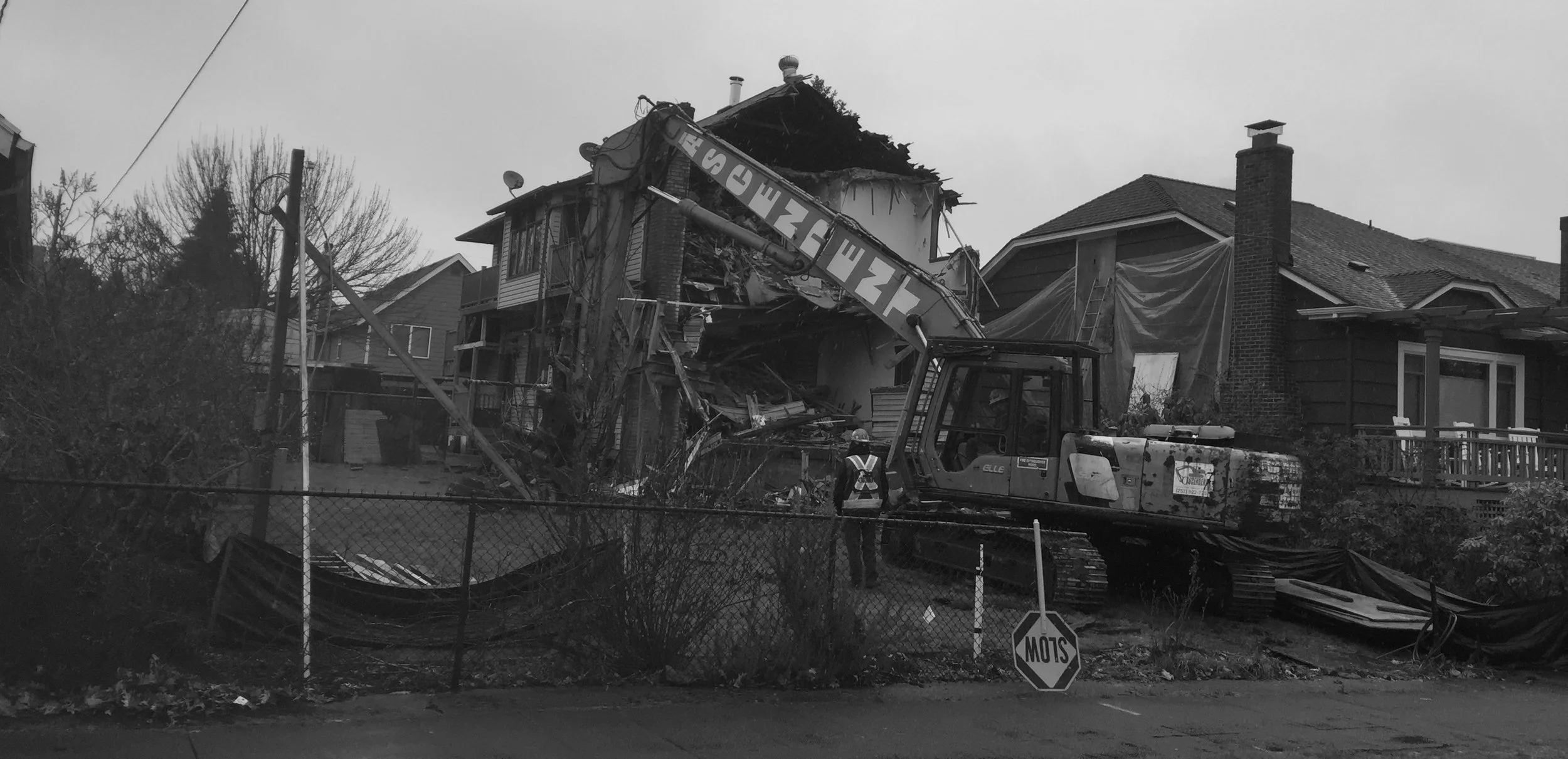 A large construction excavator demolishing a two-story house with a backyard, in a residential neighborhood, with a worker observing nearby, on an overcast day.