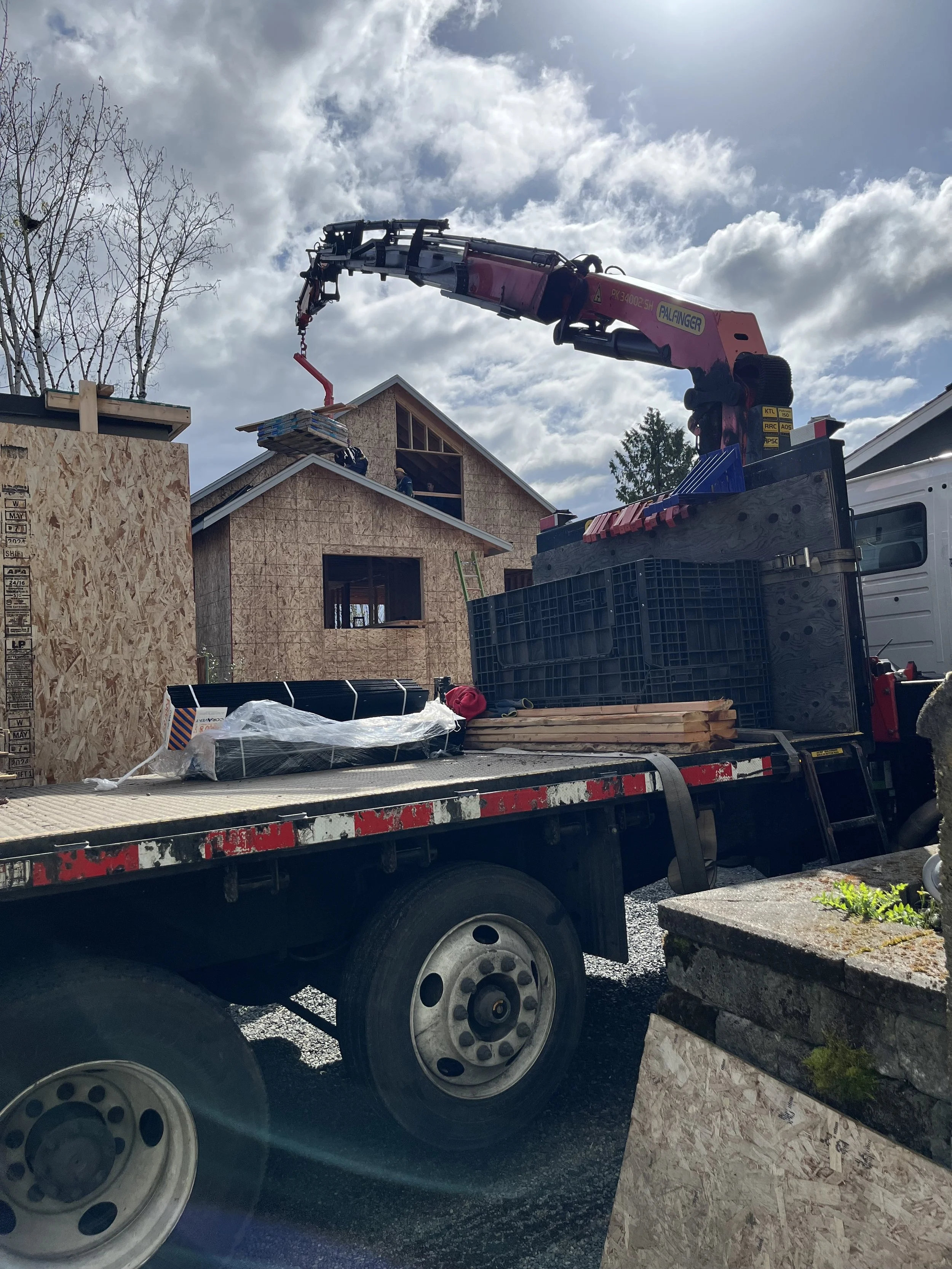Construction site with a flatbed truck and a crane lifting construction materials to a house under construction, with a partly cloudy sky.