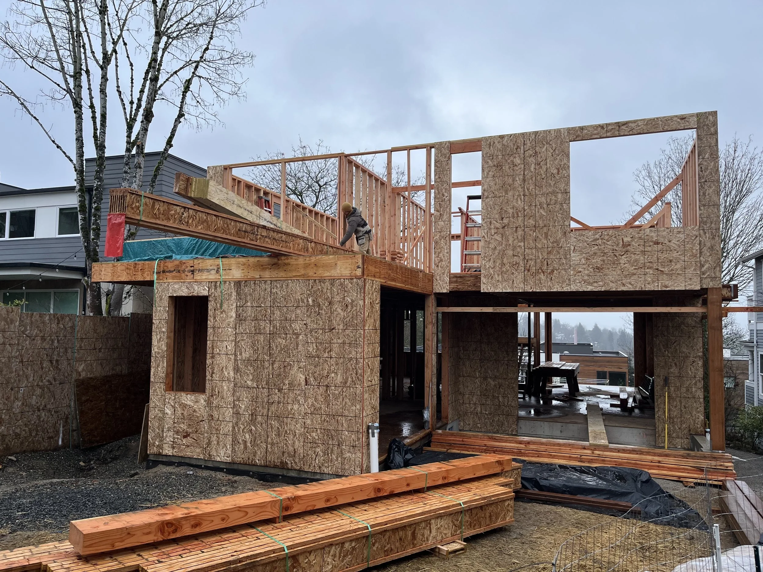 Construction site of a two-story house with wooden framing and plywood sheathing, rain-soaked ground, and a worker installing beams, surrounded by neighboring houses and leafless trees.