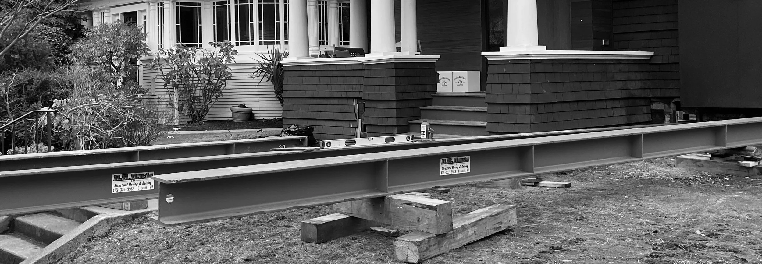 Construction site with steel beams and wooden supports in front of a house with a porch, shrubs, and stairs.