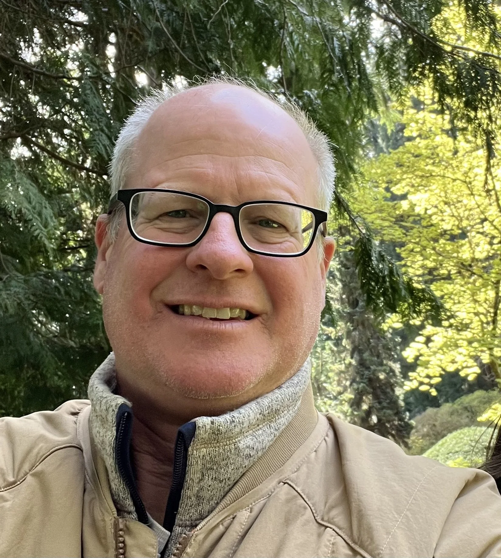 A smiling man with glasses and light-colored hair outdoors, surrounded by green trees and sunlight.