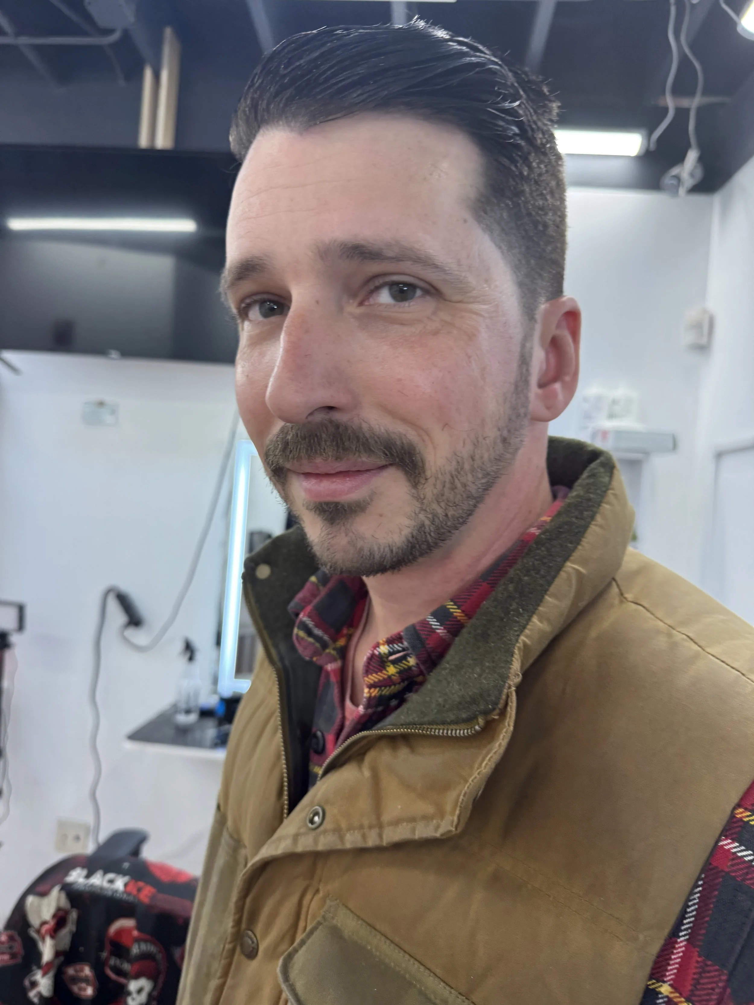 Close-up of a man with short dark hair and a beard, wearing a tan jacket over a plaid shirt, standing indoors in a room with white walls and visible wires and equipment.