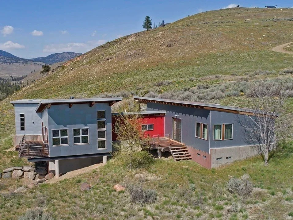 Modern house with gray and red exterior, multiple levels, and large windows in a hilly, rural landscape with grassy terrain and clear sky.