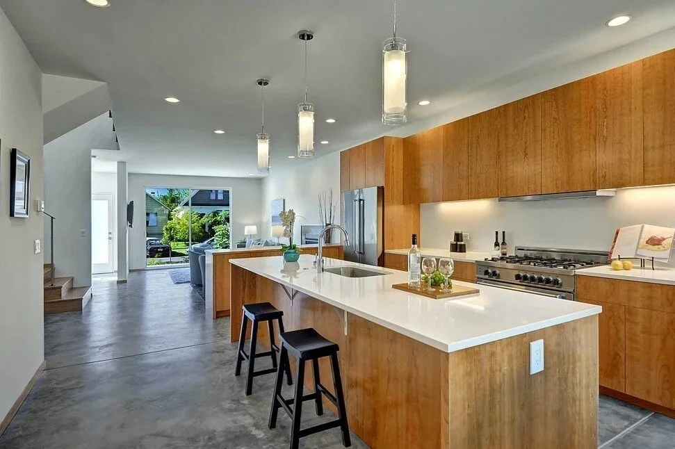Modern kitchen with wooden cabinets, white island counter, stainless steel appliances, pendant lights, and a view of the living area with large window.