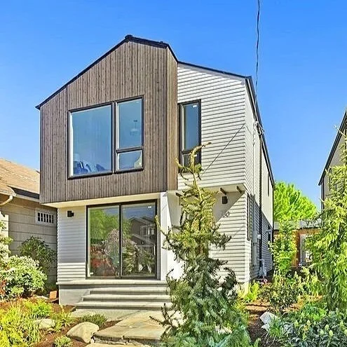 Modern two-story house with mixed siding, large front windows, and a small front porch, surrounded by garden with trees and shrubs.